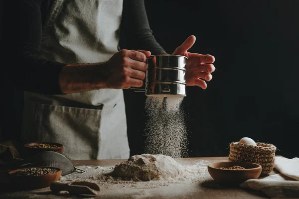 Unrecognizable young man sifting flour for kneading dough. Males hands making bread on dark background. Selective focus on the hands.