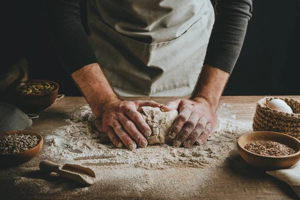 Ahşap masada hamur yoğuran tanınmaz genç adam. Erkeklerin elleri arka planda ekmek yapıyor. Seçici odak.