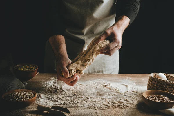Ahşap masada hamur yoğuran tanınmaz genç adam. Erkeklerin elleri arka planda ekmek yapıyor. Seçici odak.