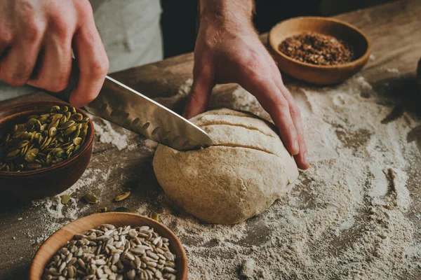 Tanımlanamayan genç adam, karanlık arka plandaki ahşap masada pişmemiş taze ekmek üzerinde bıçakla kesikler yapıyor. Seçici odak.