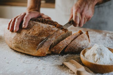 Unrecognizable young man in apron cutting homemade whole grain bread. Selective focus.