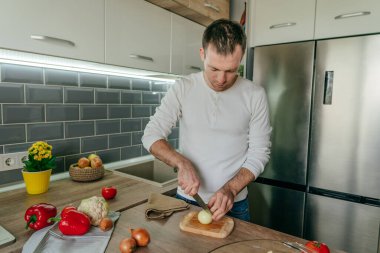 Tahtada soğan doğrayan bir adam. Genç adam mutfakta sebze pişiriyor soğan hazırlıyor. Seçici odak.