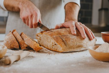 Önlüklü tanınmayan genç adam ev yapımı tahıllı ekmeği kesiyor. Seçici odak.