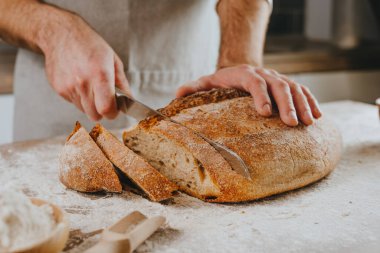 Önlüklü tanınmayan genç adam ev yapımı tahıllı ekmeği kesiyor. Seçici odak.