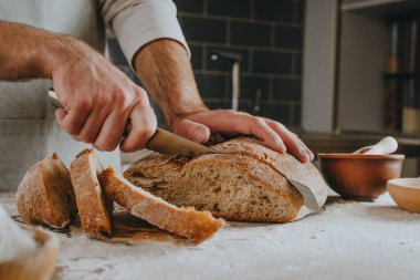 Unrecognizable young man in apron cutting homemade whole grain bread. Selective focus.