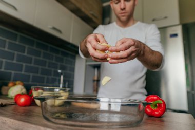 Adam tahtanın üzerinde patates kesiyor ve onu fırın tabağına koyuyor. Genç adam mutfakta sebze pişiriyor ve patates hazırlıyor. Seçici odak.