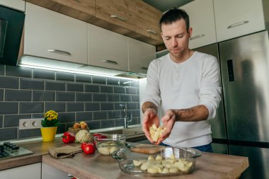 Adam tahtanın üzerinde patates kesiyor ve onu fırın tabağına koyuyor. Genç adam mutfakta sebze pişiriyor ve patates hazırlıyor. Seçici odak.