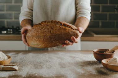 Unrecognizable young man in apron holding homemade whole grain bread. Selective focus.