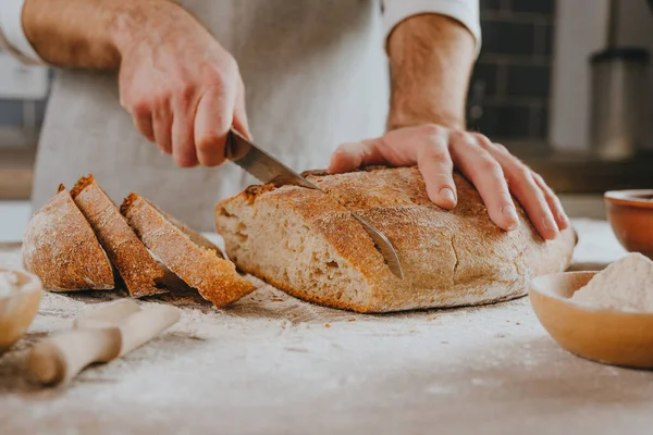 Önlüklü tanınmayan genç adam ev yapımı tahıllı ekmeği kesiyor. Seçici odak.
