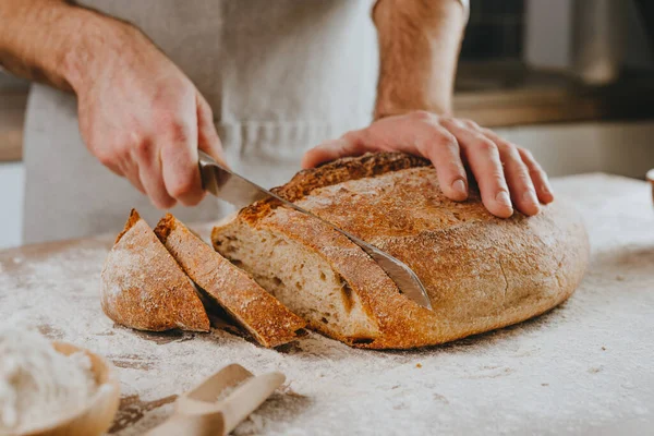 Önlüklü tanınmayan genç adam ev yapımı tahıllı ekmeği kesiyor. Seçici odak.