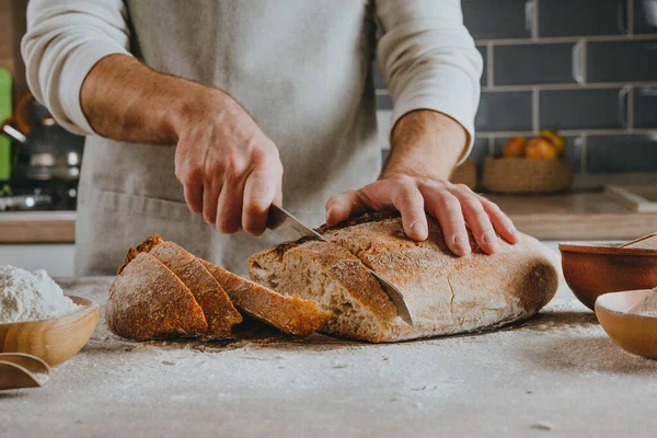 Önlüklü tanınmayan genç adam ev yapımı tahıllı ekmeği kesiyor. Seçici odak.