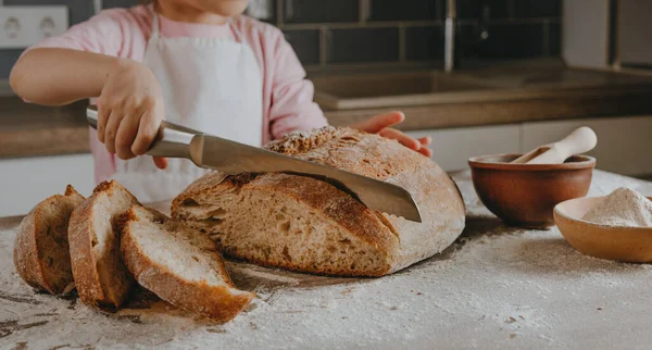 Önlüklü tanınmayan küçük kız, ev yapımı tahıl ekmeği kesiyor. Seçici odak.