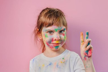 Pembe arka planda Holi renkleriyle kaplı küçük bir kızın yüzü. Cildinde renkli resimlerle gülümseyen bir bebeğin portresi..