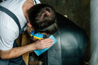 Genç bir adam zımpara kağıdıyla araba tamponunu zımparalıyor. Arabanın plastik tampon kapağını boyamadan önceki ilk adım. İşçi zararlı gaz solumamak için koruyucu ekipman kullanıyor. Seçici odak.