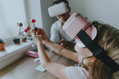 Young man describing his daughter configuration of a molecule using VR glasses. Modern technology using by family. Homeschooling concept. Selective focus.