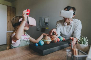Küçük kız evde güneş sistemi gezegenlerini öğrenmek için VR gözlüğü kullanarak babasıyla eğleniyor. Evde eğitim kavramı. Aile tarafından kullanılan modern teknoloji. Seçici odak.