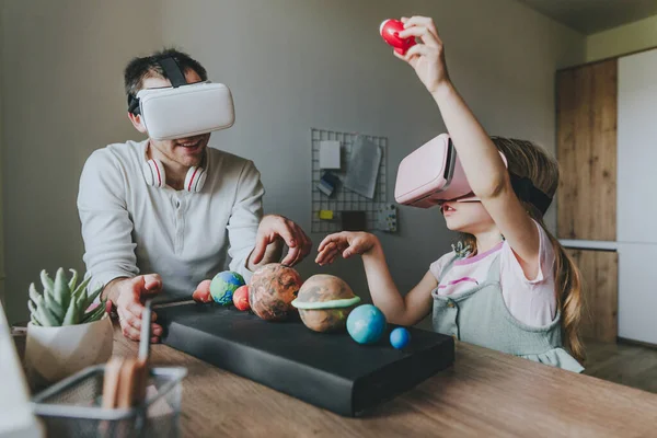Küçük kız evde güneş sistemi gezegenlerini öğrenmek için VR gözlüğü kullanarak babasıyla eğleniyor. Evde eğitim kavramı. Aile tarafından kullanılan modern teknoloji. Seçici odak.