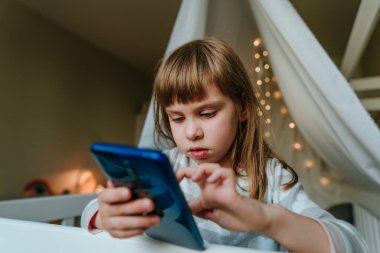 Little girl using smartphone sitting on the bed in handmade house. Generation alpha using technology. Social media concept.