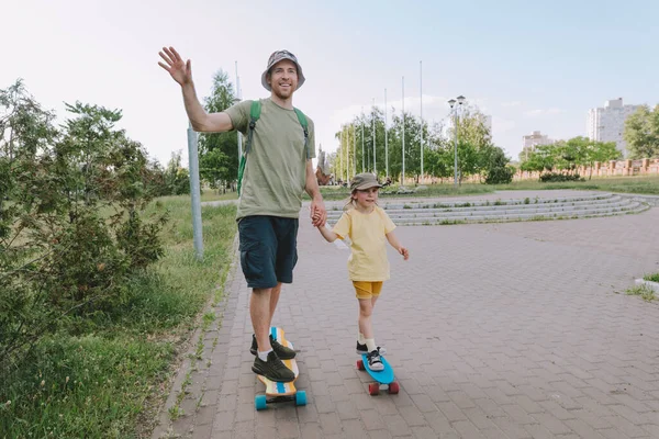 Happy father and his daughter riding on skateboards outdoor. Young man and preschooler girl enjoying having fun time together at summer. Family activities concept.