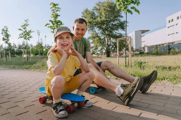 Happy father and his daughter riding sitting on skateboards outdoor. Playful young man and preschooler girl enjoying having fun time together at summer. Family activities concept.