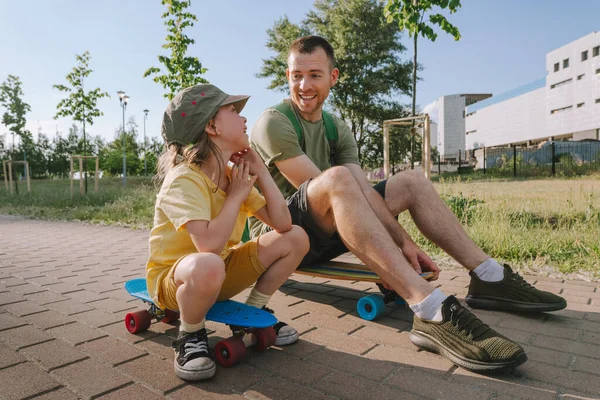 Happy father and his daughter riding sitting on skateboards outdoor. Playful young man and preschooler girl enjoying having fun time together at summer. Family activities concept.