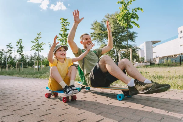 Happy father and his daughter riding sitting on skateboards outdoor. Playful young man and preschooler girl enjoying having fun time together at summer. Family activities concept.