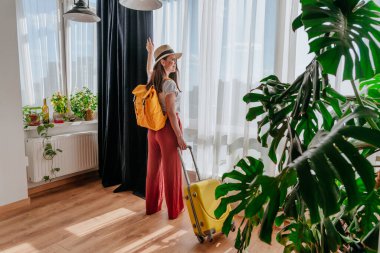 Pencereden bakan genç bir kadının yeni şehir manzarasının tadını çıkarışının arka görüntüsü. Otel odasına çanta ve sırt çantalı bir turist geliyor..