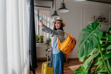 Genç kadın blogcu, bir otel odasında tatilin tadını çıkarırken akıllı telefon kullanarak selfie çekiyor. Çantalı ve sırt çantalı turist çevrimiçi oda turu yapıyor..