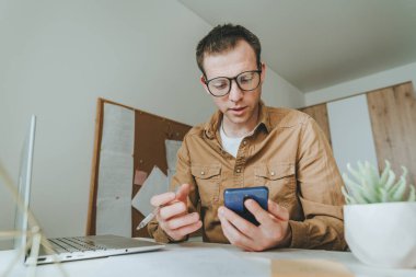 Genç adam odada masada otururken görüntülü konuşma için akıllı telefon ve dizüstü bilgisayar kullanıyor. Serbest Çalışan çevrimiçi konferans veriyor. Evdeki teknolojiyi kullanarak. Seçici odak.