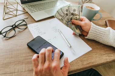 Akıllı telefondaki uygulamayı kullanarak ve el yazısıyla hesaplama yaparak Amerikan dolarlarını elinde tutan genç adam bütçesini kapat. Seçici odak.