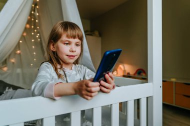 Little girl using smartphone sitting on the bed in handmade house. Generation alpha using technology. Social media concept.