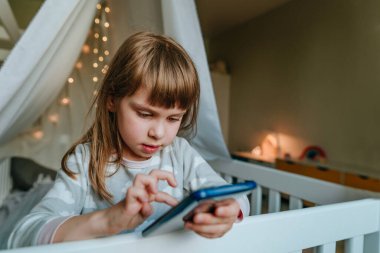 Little girl using smartphone sitting on the bed in handmade house. Generation alpha using technology. Social media concept.