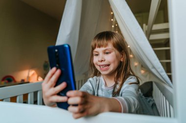 Little girl using smartphone sitting on the bed in handmade house. Generation alpha using technology. Social media concept.