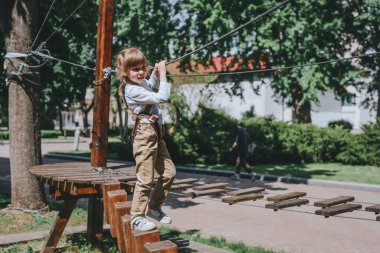 Anaokulundaki küçük kız tırmanma takımı takıyor güvenlik ekipmanlarıyla ip parkında eğleniyor. Çocuklar için yaz kampı aktivitesi. Ormandaki macera parkı.