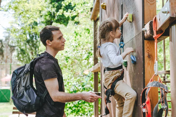 Young father teaching her daughter to climb on the rock climbing wall. Little girl preschooler wearing safety harness having fun time in adventure rope park. Happy family concept.