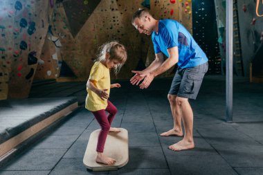 Babası, spor salonunda spor yaparken kızına destek oluyor. Küçük kız spor aksesuarları kullanıyor. Aile spor aktiviteleri. Arka planda kaya tırmanışı duvarı.