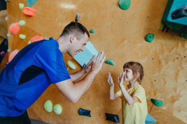 Babası ve kızı kaya tırmanışçıları modern kapalı bir kaya tırmanış salonunda eğitim görüyorlar. Erkek eğitmen özel bir koç sınıfında küçük kız anaokuluna beşlik çakıyor. Aile spor aktiviteleri. Seçici odak.