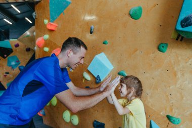 Babası ve kızı kaya tırmanışçıları modern kapalı bir kaya tırmanış salonunda eğitim görüyorlar. Erkek eğitmen özel bir koç sınıfında küçük kız anaokuluna beşlik çakıyor. Aile spor aktiviteleri. Seçici odak.
