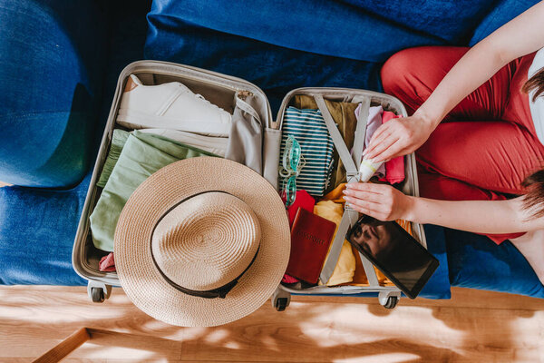 Вид сверху красивой молодой женщины, упаковывающей личные вещи в чемодан для путешествий, глядя на список за столиком, сидящим на диване в гостиной.