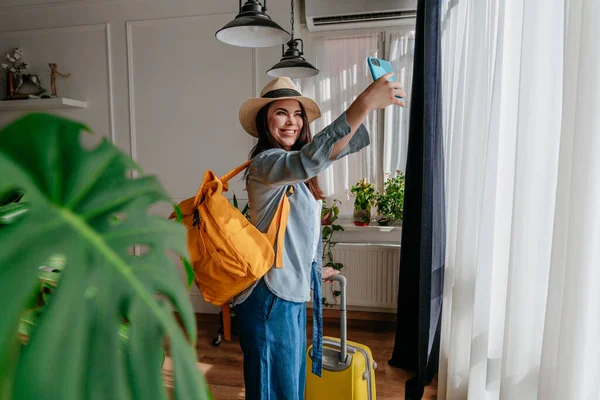 Young woman blogger making selfie using smartphone in a hotel room enjoying her vacation. Tourist with suitcase and backpack making online room tour.