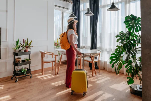 Pencereden bakan genç bir kadının yeni şehir manzarasının tadını çıkarışının arka görüntüsü. Otel odasına çanta ve sırt çantalı bir turist geliyor..
