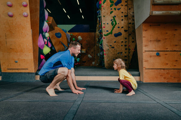 Father and daughter doing squats in a gym. Family sport activities. Rock climbing wall on background.