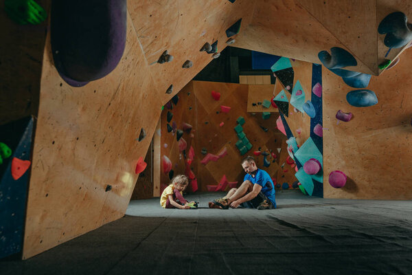 Father and his daughter rock climbers putting on climbing shoes in a modern climbing wall gym sitting on the floor. Instructor teaching little girl preschooler how to climbing. Family sport activities. Selective focus.