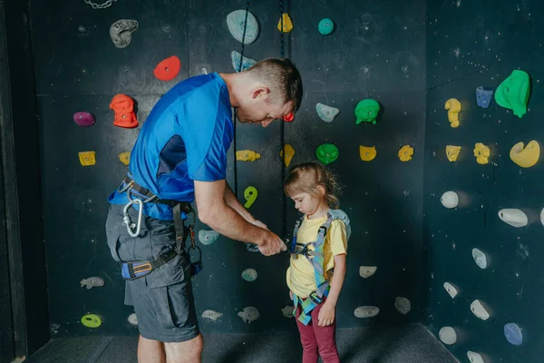 Father and his daughter rock climbers training in a modern indoor rock climbing gym. Male instructor helps little girl preschooler putting on safety harnesses on a coach class.