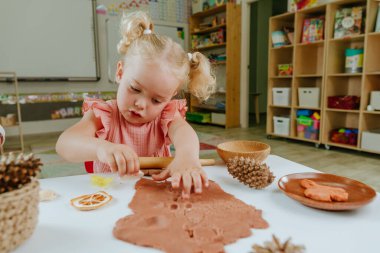 Anaokulundaki kız hamura doğal malzemeler kullanarak parmak izi yapıyor. Portakal dilimleri, koniler, kabuklar, anaokulundaki tüyler. Erken kalkınma kavramı.
