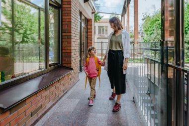 Mother leading her daughter at school. Young woman and little 6 years old girl near elementary school building. Back to school concept.