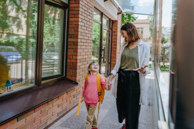 Mother leading her daughter at school. Young woman and little 6 years old girl near elementary school building. Back to school concept.
