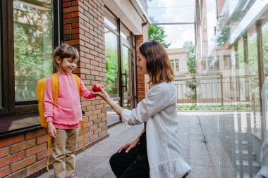 Mother leading her daughter at school giving an apple. Young woman and little 6 years old girl near elementary school building. Back to school concept.