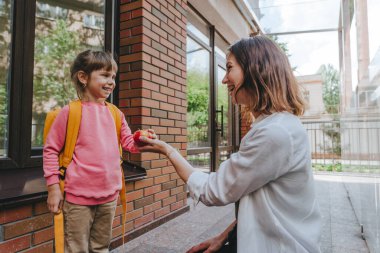 Mother leading her daughter at school giving an apple. Young woman and little 6 years old girl near elementary school building. Back to school concept.