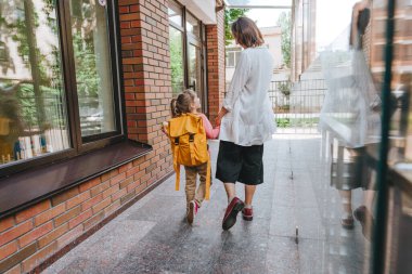 Mother leading her daughter at school. Back view of young woman and little 6 years old girl near elementary school building. Back to school concept.
